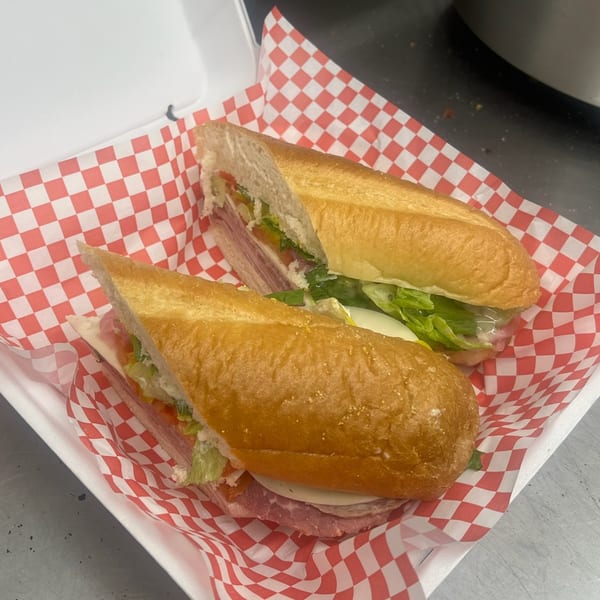 Two Mia Ava East Coast Pizza subs, cut in half, in a white container with red and white checkered paper.
